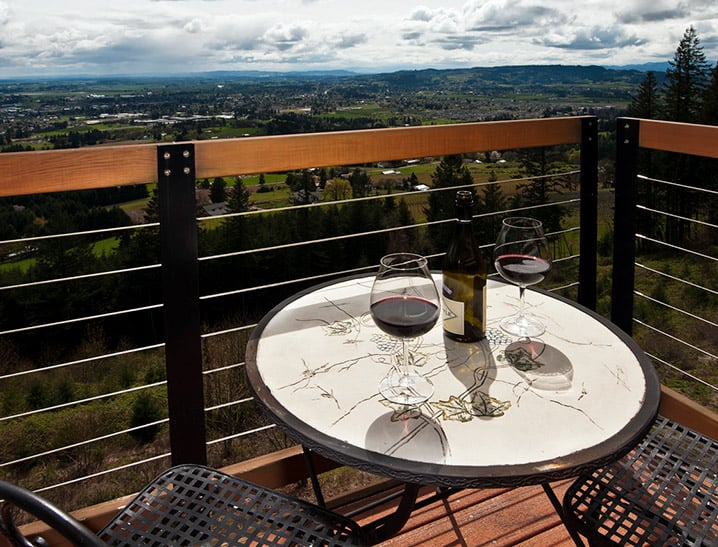 A table and chairs with wine glasses on a deck overlooking rolling hills and blue skies, in Newberg, Oregon.
