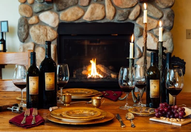 A table set with plates, silverware, wine glasses, candles and in front of a fireplace.