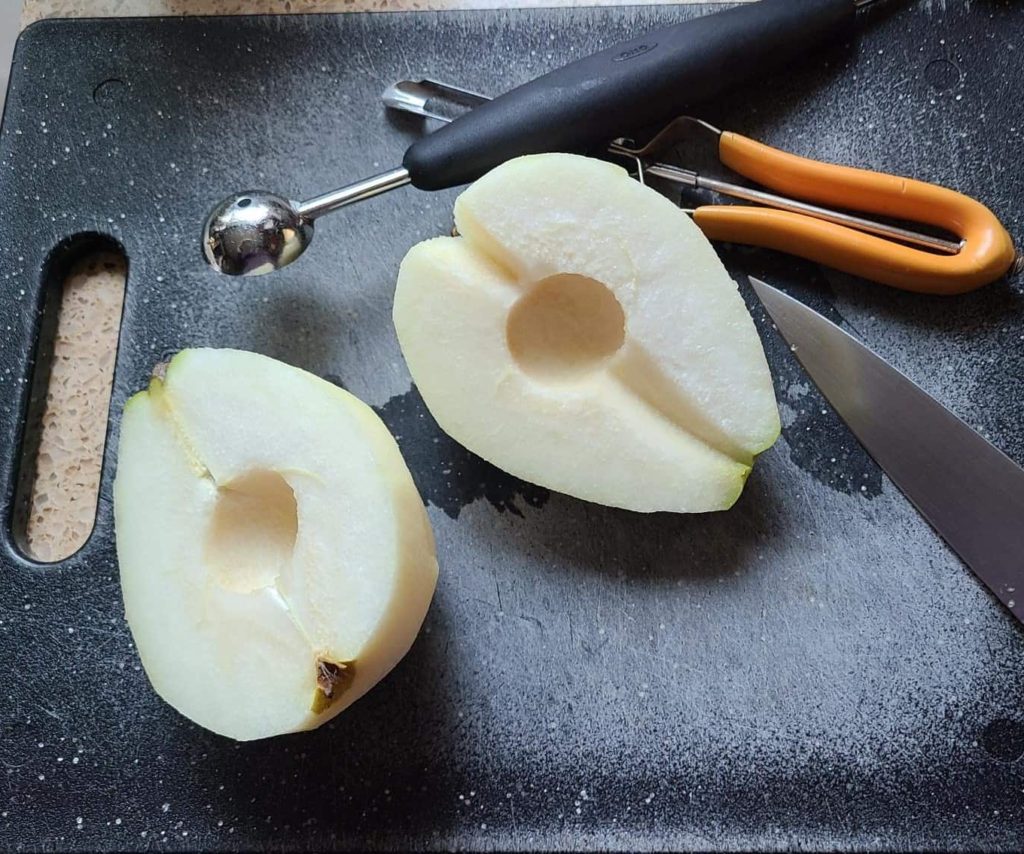 two pear halves, with the core cut out on a cutting board with a peeler and melon baller