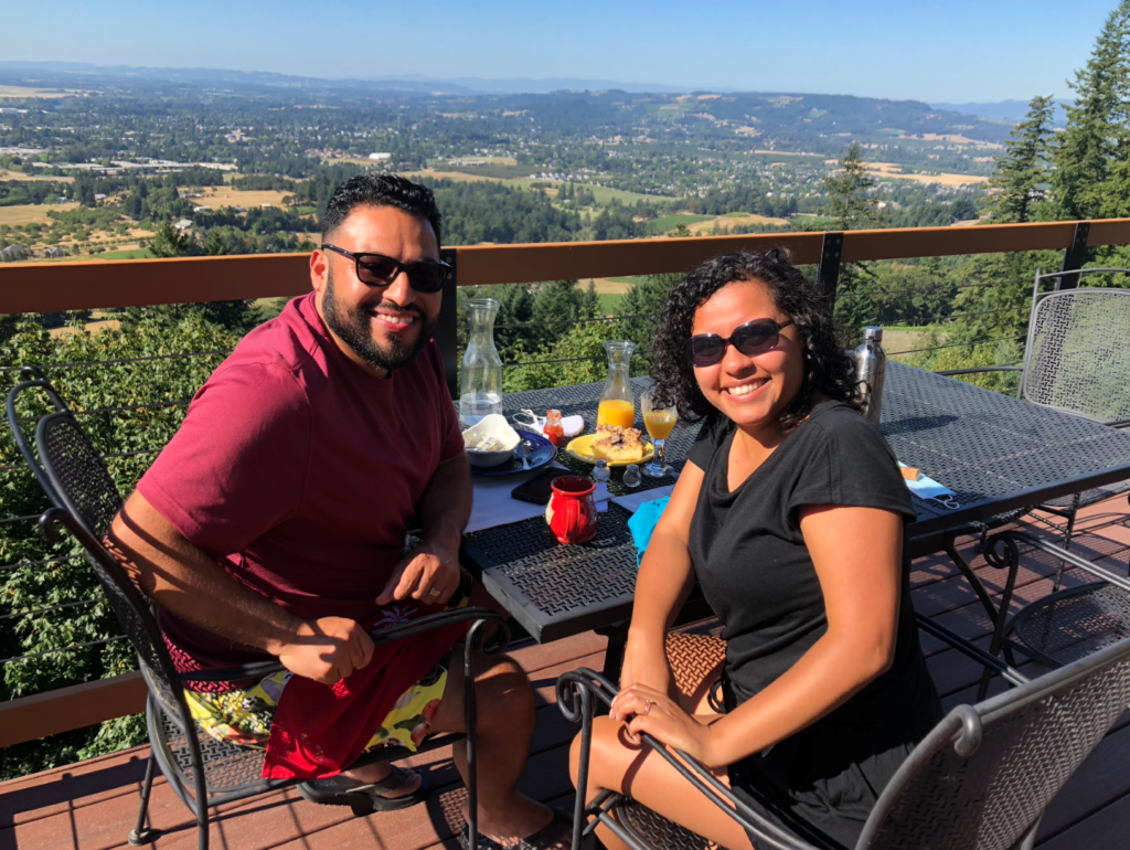 a man and a woman sitting at a breakfast table on a deck overlooking a valley