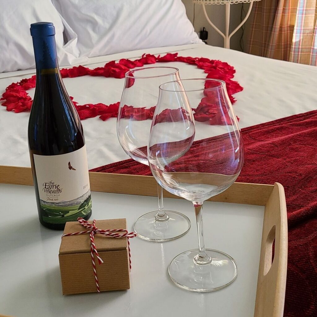 a bed with flower petals on it in a heart shape, with a bottle of wine and a vase of red roses