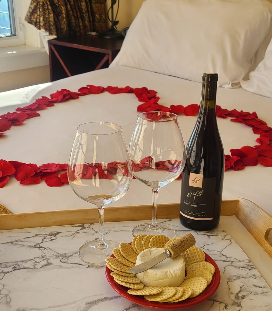 Truffle Love Package, rose petals on a bed, bottle of wine with wine glasses and a plate with spreadable cheese and truffle crackers