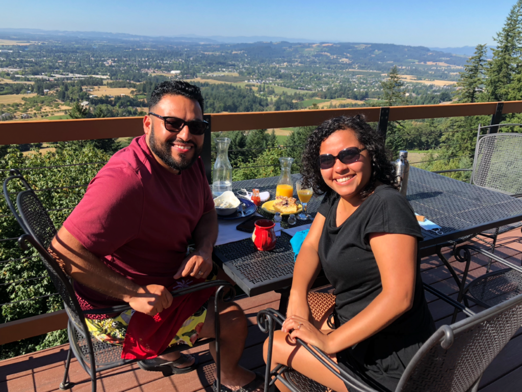 A man and a woman sitting at an outside table, looking at the camera with a view of the Willamette Valley in the background and breakfast set on the table