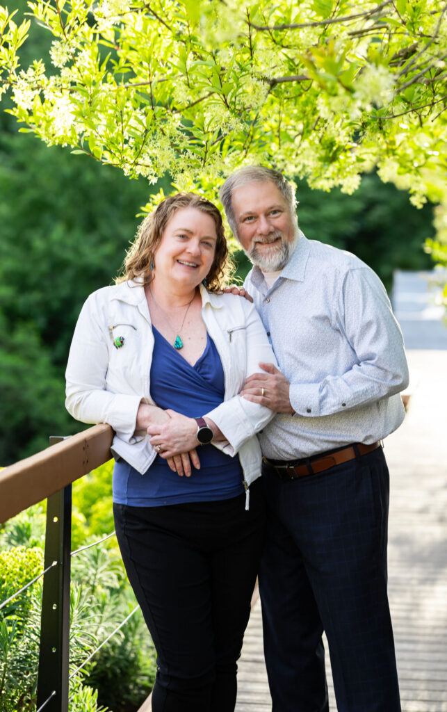 a woman and man standing together against a deck railing, with tree foliage in the background