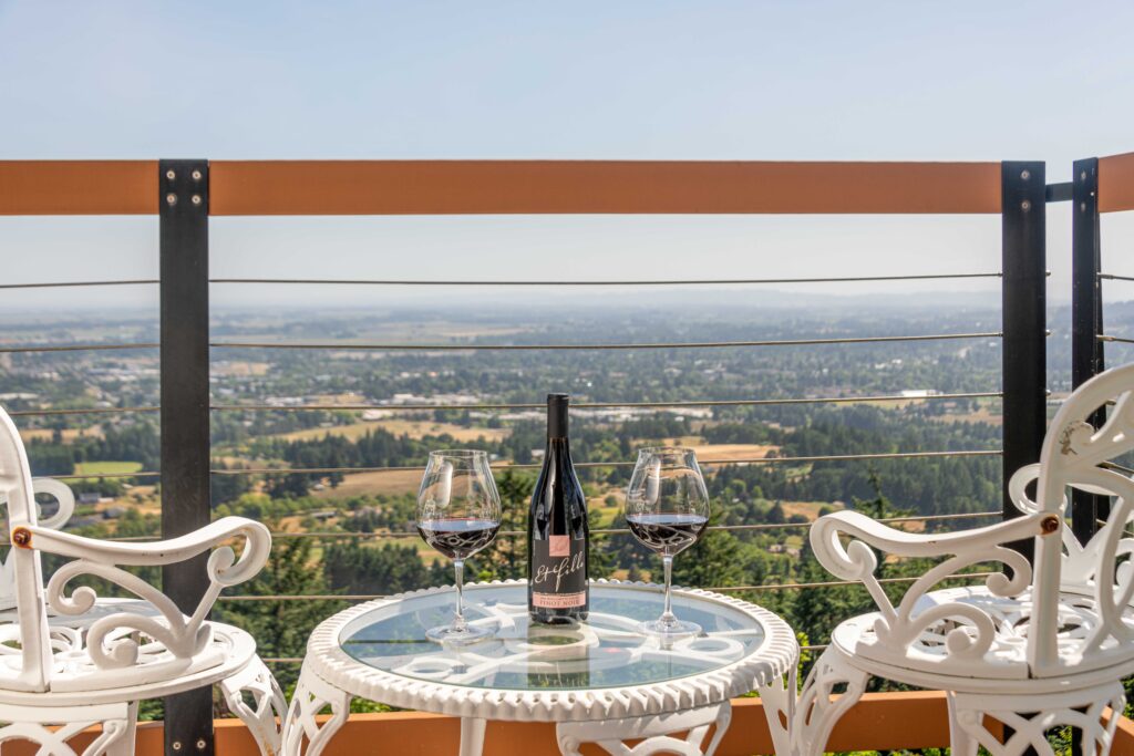 Scenic terrace view with a glass-top table and two white ornate chairs overlooking a lush valley. On the table sit two glasses of red wine and a bottle of Et Fille Pinot Noir, framed by a modern railing and a clear blue sky and views of the willamette valley