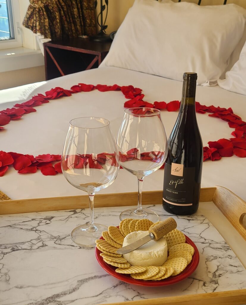 A romantic hotel room setup with a white bed decorated with red rose petals arranged in a heart shape. In the foreground, a tray holds two empty wine glasses, a bottle of red wine, and a small round of cheese surrounded by crackers with a cheese knife.