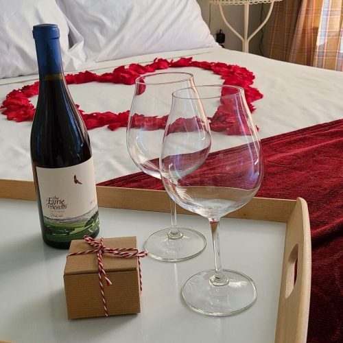 a bed with flower petals on it in a heart shape, with a bottle of wine and a vase of red roses