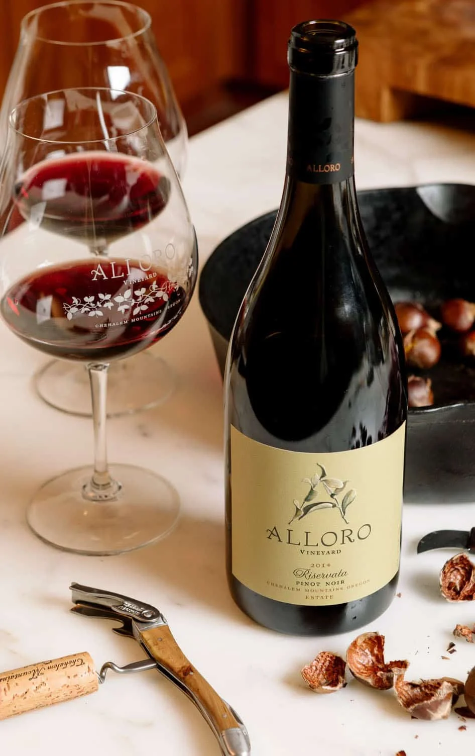 a bottle of wine, with 2 pinot noir glasses beside it, both with about 2 ounces of wine, cracked and whole hazelnuts on the table